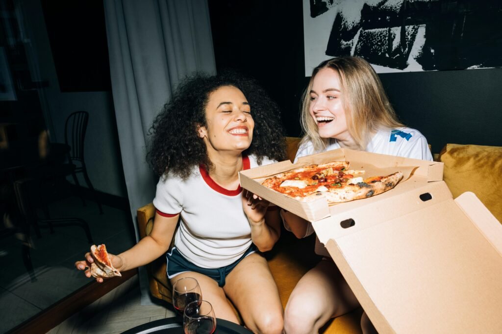 Two happy friends laughing and enjoying pizza indoors during a cozy night in.